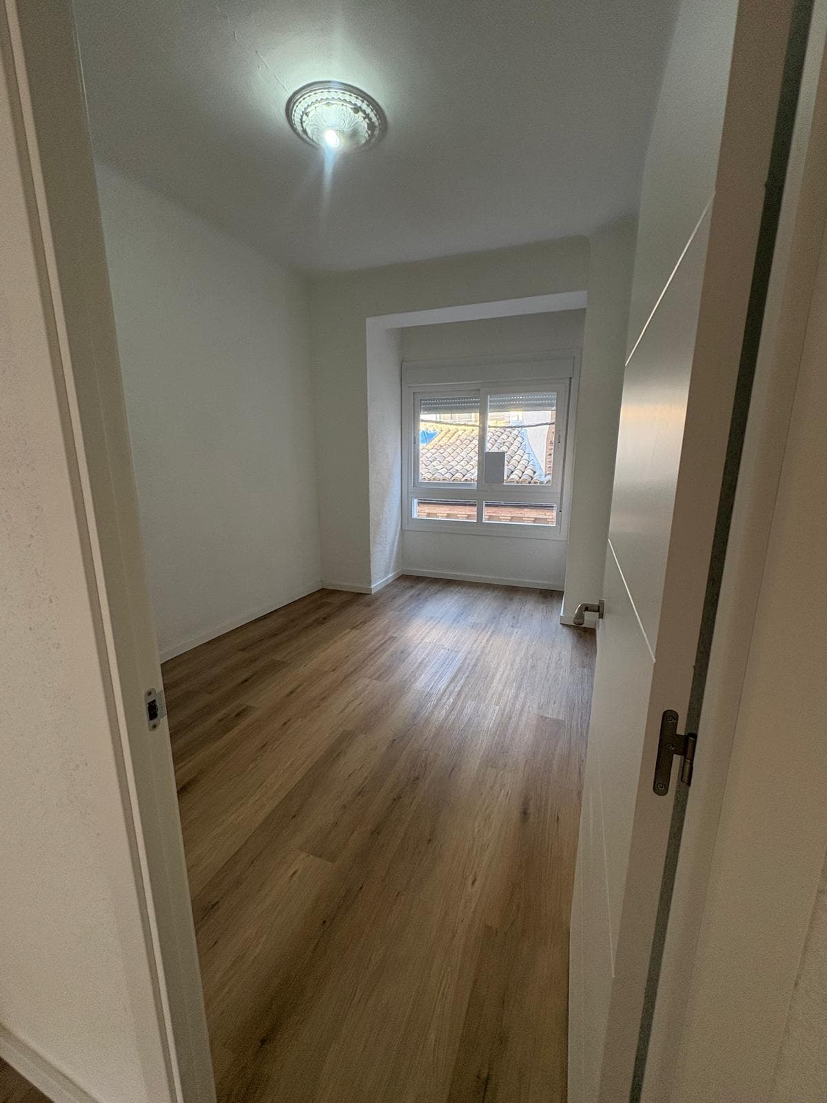 Habitación reformada con suelo laminado, paredes blancas y ventana nueva, lista para su siguiente uso